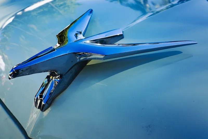 Close Up of a Chrome Bird Hood Ornament on a Light Blue Classic Car - The image shows a chrome bird hood ornament, positioned on the front of a light blue classic car. The ornament is highly polished and reflects the light of the surroundings. The bird is in flight, with wings spread, and has a long, slender beak. The car's paint is also glossy, and the background is blurred, making the hood ornament the focal point of the image.